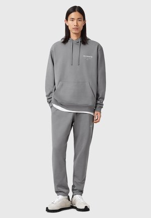 Young person standing straight wearing gray hoodie and matching joggers with white sneakers against plain background.