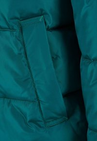 Tkanina kurtki puchowej w kolorze morskim z pikowanym wzorem, wyposażona w boczną kieszeń z klapą i kontrastującym przeszyciem.