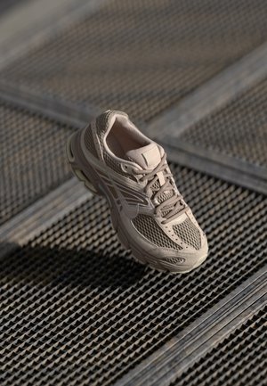 Béžová atletická teniska z síťoviny a kůže se šněrováním na texturovaném kovovém roštu, osvětlená přirozeným světlem vrhajícím stíny.