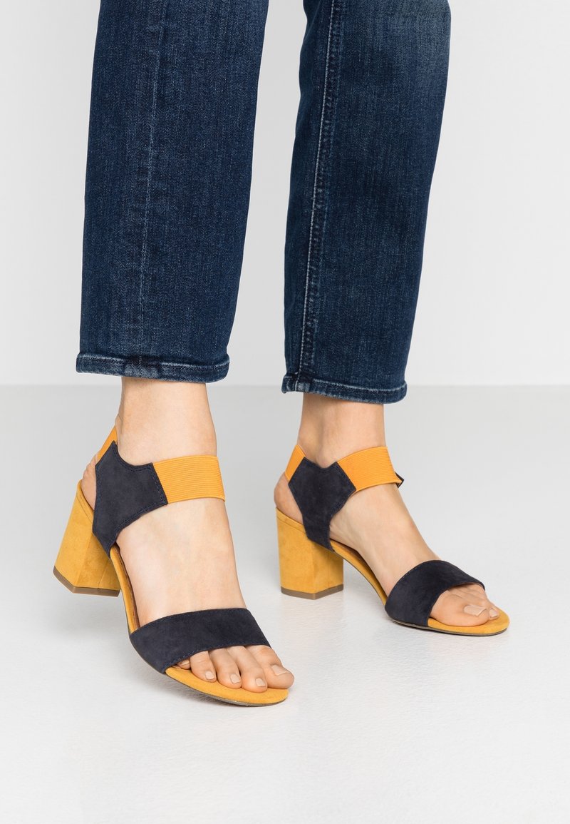 Pieds portant des sandales à talons blocs avec des brides noires et orange, associées à un jean bleu foncé retroussé sur un sol blanc.