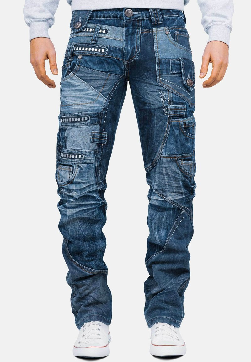 Pantaloni cargo in denim blu scuro con multiple tasche, cuciture a contrasto e una texture leggermente rovinata. Presentano chiusure a bottone e cerniera.