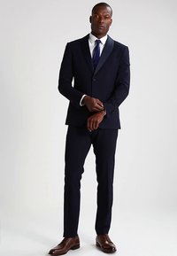 Tumesinine ülikond ühe rinnaga bleiseri ja sälguga kraega, kombineeritud sobivate pükstega ja valge kleidisärgiga, mida täiendab sinine lips.