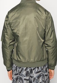 Olive green bomber jacket in smooth, shiny fabric with ribbed collar and cuffs. Features a side pocket and elasticized waistband.