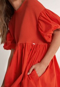 Vestido de textura roja con mangas cortas abullonadas, cintura fruncida y bolsillos laterales. Presenta una parte inferior de tela suave con un detalle sutil de etiqueta.