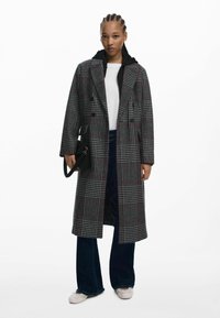 Abrigo de cuadros largo en gris con acentos rojos, diseño de doble botonadura, solapas en pico y bolsillos laterales; lo lleva puesto sobre una sudadera negra con capucha y unos vaqueros azules.