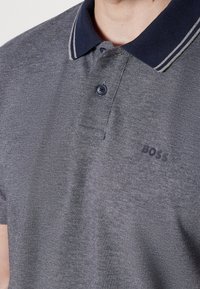 Polo gris texturé avec col bleu marine et deux rayures blanches, boutonné, avec le logo "BOSS" sur la poitrine gauche.
