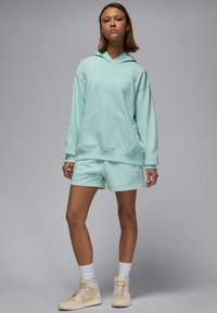 Sudadera de color verde menta claro con bolsillo tipo canguro, combinada con pantalones cortos a juego. Logo de acento en el pecho. Llevada con zapatillas altas color crema.