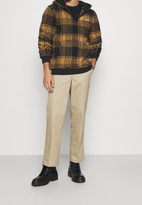 Pull polaire à carreaux noir et jaune avec un col à demi-fermeture éclair, associé à un pantalon beige et des bottes noires à lacets.