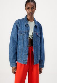 Denimjakke med et teksturert blått mønster, frontlommer, metallknapper og en avslappet passform, kombinert med røde bukser med snøring.
