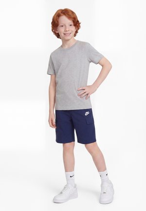 Băiat roșcat zâmbitor stă cu mâna pe șold, purtând tricou gri, pantaloni scurți bleumarin cu logo Nike, șosete albe Nike și pantofi sport albi.