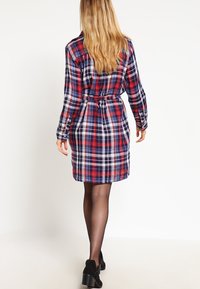 Robe chemise à carreaux aux tons rouge, blanc et bleu, devant boutonné, manches longues, ceinture ajustable à la taille, et chaussures solides noires.