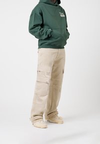 Sweat à capuche vert avec poche avant, pantalon cargo beige avec plusieurs poches, et baskets beiges à brides. Design simple et coupe décontractée.