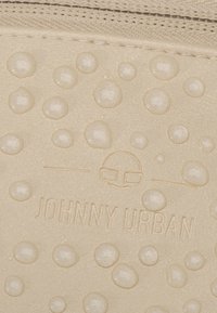 Gouttes d'eau sur un tissu beige avec le logo de la marque "Johnny Urban" en relief et un visage stylisé portant des lunettes au-dessus du texte.