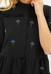 Abito nero con maniche corte, colletto arruffato, chiusura a bottoni e accenti floreali ricamati in verde su fondo blu. Gonna leggermente arricciata.