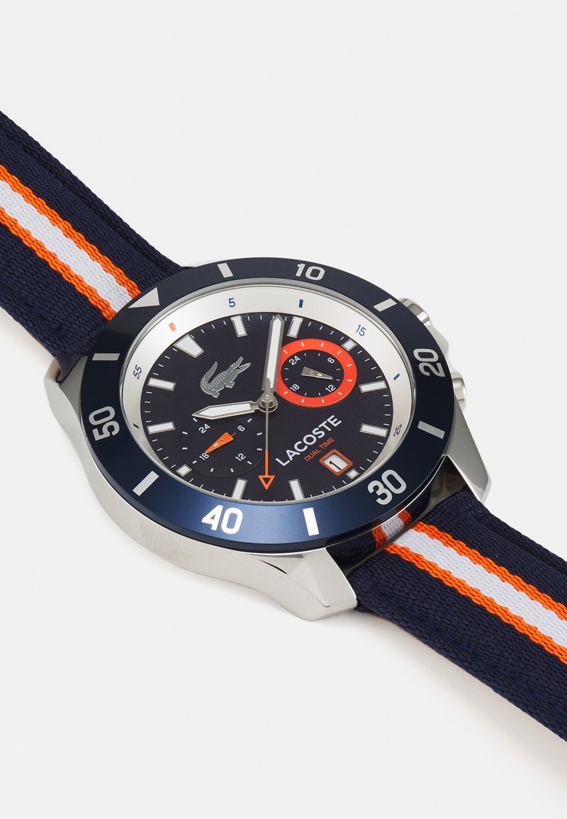 Reloj de marina con acentos plateados, una gran caja redonda, una correa de tela a rayas con líneas naranjas y blancas, y una esfera negra con marcadores luminiscentes.