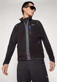 Man in een zwart fleece vest met blauwe ritssluitingen, een zwart shirt met lange mouwen, zwarte broek en grote zwarte zonnebril, poserend tegen een effen lichte achtergrond.
