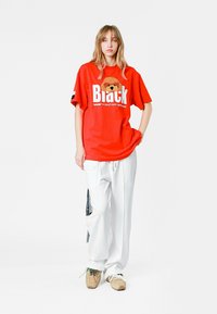 Camiseta roja de gran tamaño con un gráfico de un oso y el texto "Black", combinada con pantalones blancos de corte holgado con detalles laterales y zapatillas beiges.