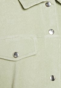 Tessuto di velluto a coste verde chiaro con righe verticali, caratterizzato da chiodini metallici e una tasca a patta. La texture è morbida e liscia.