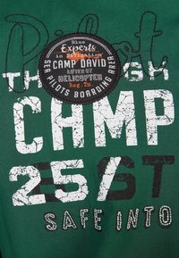 T-shirt en coton vert avec texte blanc usé, un écusson rond aux détails noirs et orange, et des accents de couture contrastants.