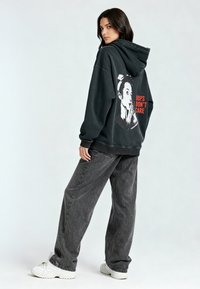 Fekete, nagy méretű kapucnis pulóver, melynek hátoldalán egy arcot ábrázoló grafikát és piros színű "Oops I Don’t Care" feliratot találunk. Szürke farmerrel és fehér tornacipővel párosítva.