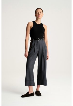 Kobieta stojąca, ubrana w czarny top bez rękawów, szare szerokie spodnie z wysokim stanem i paskiem oraz czarne wsuwane buty na jednolitym tle.