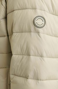 Veste doudoune beige clair avec un design matelassé. Présente un badge logo circulaire avec du texte. Texture lisse avec un léger brillant.
