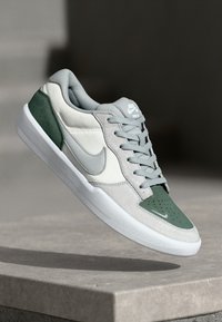 Sneakers Nike cu un amestec de materiale gri, alb și verde. Prezintă o parte superioară netedă, vârf perforat și accente texturate.