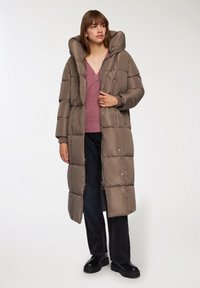 Giacca puffa color taupe con colletto alto, zip frontale e polsini a coste. Abbinate a un maglione rosa e jeans neri a gamba larga. Scarpe nere.