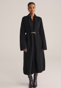 Sieviete, kas valkā garu melnu kardigānu ar zelta sprādzi, melnu kleitu, auskariem ar riņķiem un tumši brūnām augstām zābakiem.