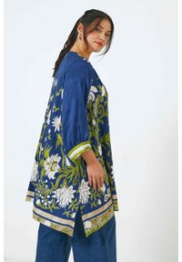 Blauwe katoenen tuniek met witte en groene bloemenpatronen, driekwart mouwen en gestreepte groene accenten op de zoom en mouwen.