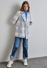 Nainen, jolla on päällään sininen pooloneule, kermanvärinen ja sinivalkoinen ruudullinen takki, käärityt farkut ja valkoiset tennarit, seisoo vaaleanharmaata seinää vasten.