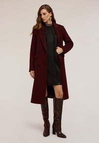 Cappotto in mistura di lana bordeaux con design doppiopetto, bottoni neri e tasche laterali. Indossato sopra un vestito nero con trama e stivali in pelle di serpente.