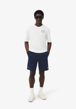 Uomo con occhiali da sole, t-shirt bianca Lacoste, pantaloncini blu navy, calzini bianchi e sneakers beige, in piedi con le mani in tasca.