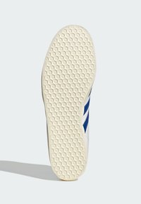 Bijela tenisica s plavim prugama, koja ima teksturiranu gumenu podlogu s malim uzorcima zvjezdica i glatki gornji dizajn.