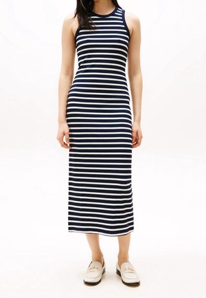 Femme portant une robe midi sans manches à rayures horizontales bleu marine et blanches, avec des mocassins crème, sur un fond blanc uni.