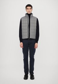 Gilet grigio trapuntato con colletto alto, chiusura lampo sul davanti, indossato sopra un dolcevita nero, abbinato a pantaloni blu navy e scarpe nere. Sfondo semplice.