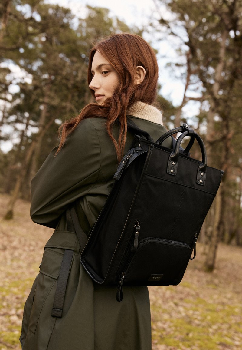 Black fabric backpack with dual handles, a front zip pocket, and leather accents. Features a structured shape and adjustable straps.