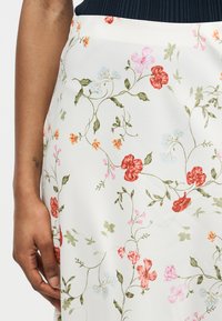 Κοντινό πλάνο ενός χεριού που ακουμπά δίπλα σε μια λευκή φούστα με πολύχρωμο μοτίβο από λουλουδάκια και σκούρο μπλε λάστιχο μέσης.