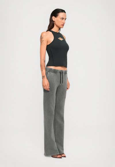 Woman with wet hair wearing black sleeveless top with chest cut-out, gray drawstring trousers, and brown open-toe heels, standing in profile.