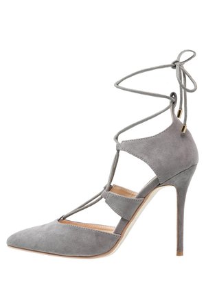 Grijze suède pumps met hoge hak, puntige neus, uitgesneden zijkanten en enkellinten die rondom de enkel zijn gewikkeld en aan de achterkant in een strik zijn gebonden.