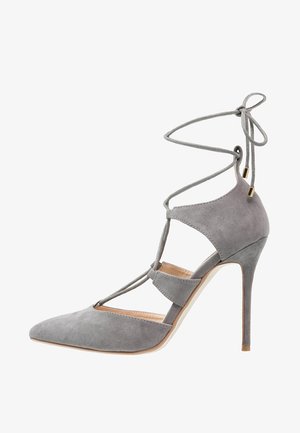 Zapato de tacón alto de ante gris con punta puntiaguda, lados recortados y cordones alrededor del tobillo atados en un lazo en la parte posterior.