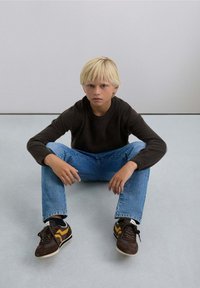 Un garçon assis sur le sol, portant un t-shirt à manches longues gris foncé, un jean bleu et des baskets marron avec des accents jaunes. Fond lisse.