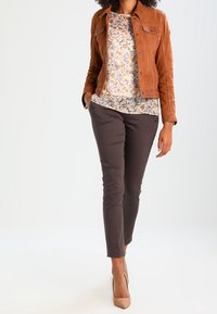 Giacca in pelle scamosciata marrone sopra una blusa a fantasia con foglie autunnali. Pantaloni marroni aderenti e décolleté beige chiaro completano l'outfit.