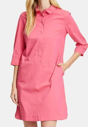 Frau trägt ein korallenrotes Kleid mit Kragen und dreiviertellangen Ärmeln, eine Hand in der seitlichen Tasche, vor weißem Hintergrund.