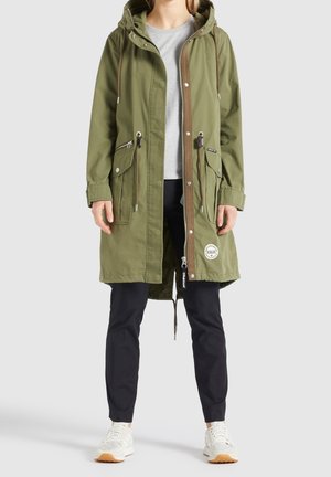 Persoon die een groene gewatteerde parka met capuchon draagt, met zakken aan de voorkant en een witte ronde patch, over een grijs shirt en zwarte broek, staand.