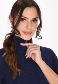 Marineblå fleece-pullover med halv-lynlås krave, der har en blød tekstur. Modellen har langt, bølget hår og en neutral makeup.