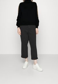 Suéter negro de cuello alto con cinta de logo en las mangas, combinado con pantalones oscuros con dobladillo vuelto y zapatillas de plataforma blancas, todo sobre un fondo neutro.