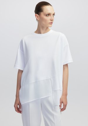 T-shirt blanc oversize en tissu lisse avec des manches courtes, doté d'un design superposé et d'un ourlet incurvé, associé à un pantalon blanc.