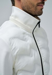 White puffer jacket with a high collar, featuring a black zipper and quilted texture. Smooth fabric with subtle sheen and minimalistic design.