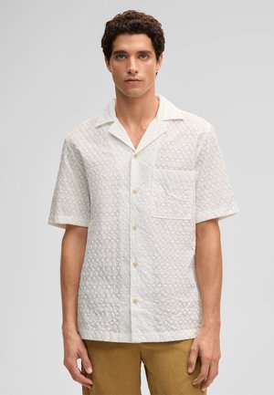 Junger Mann mit lockigem dunklem Haar, der ein kurzärmeliges, weißes, strukturiertes Hemd mit Knöpfen und beige Hosen trägt, steht vor einem schlichten Hintergrund.